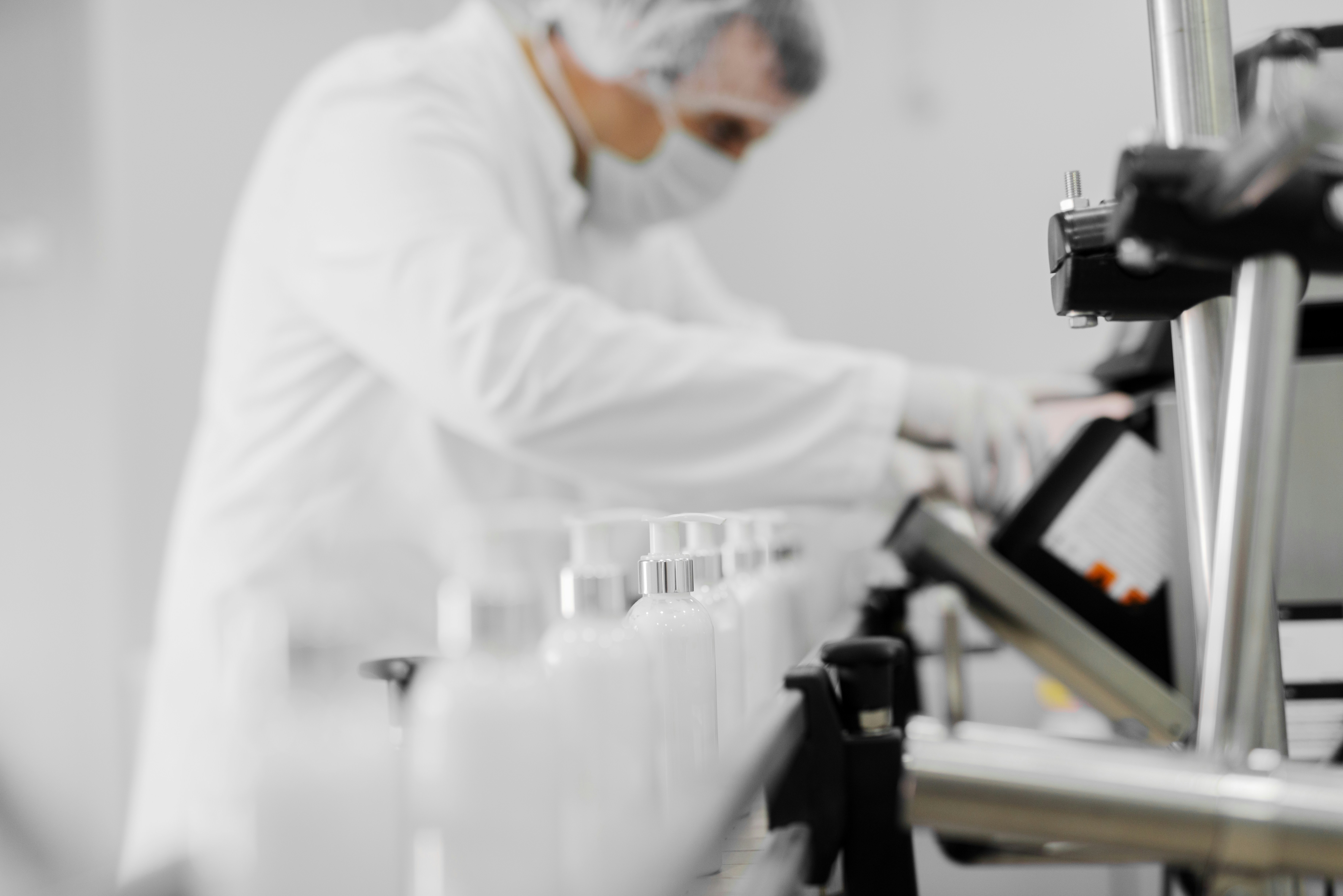 White pump bottles moving along a packaging conveyor while a worker in a cleanroom gown and mask tends the line
